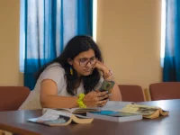 Mahasiswi berkacamata menatap ponsel di meja perpustakaan dengan buku-buku terbuka di depannya