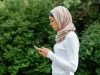 Ilustrasi seorang perempuan berjilbab menatap layar telepon pintar sambil berdiri di depan rimbun dedaunan hijau.
