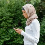 Ilustrasi seorang perempuan berjilbab menatap layar telepon pintar sambil berdiri di depan rimbun dedaunan hijau.