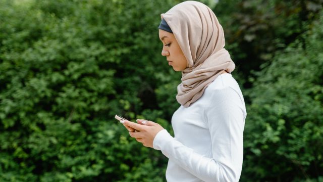 Ilustrasi seorang perempuan berjilbab menatap layar telepon pintar sambil berdiri di depan rimbun dedaunan hijau.