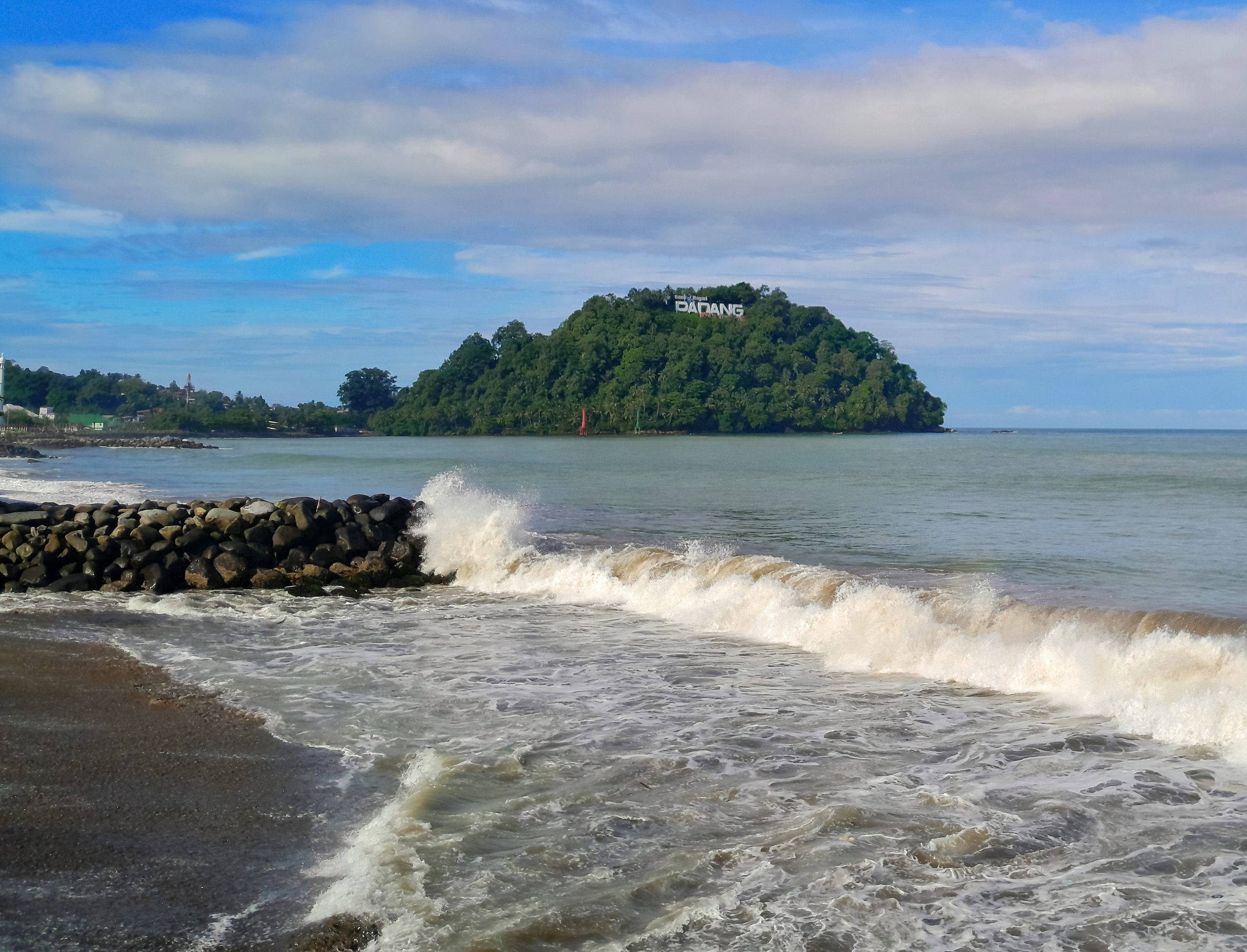 Ombak memecah di Pantai Padang dengan bukit hijau Gunung Padang di kejauhan.