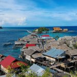 Ilustrasi permukiman rumah panggung beratap seng berdesakan di tepi laut biru di antara batu karang dan pepohonan.
