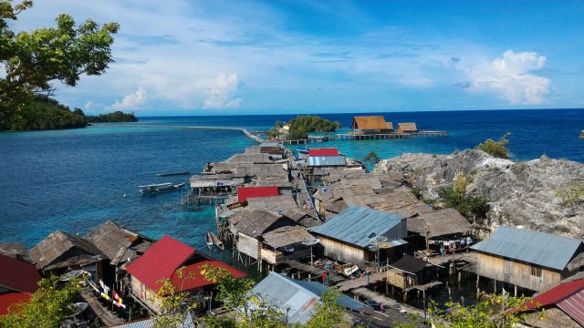 Ilustrasi permukiman rumah panggung beratap seng berdesakan di tepi laut biru di antara batu karang dan pepohonan.