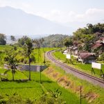 Ilustrasi rel kereta melengkung membelah hamparan sawah hijau dan permukiman.