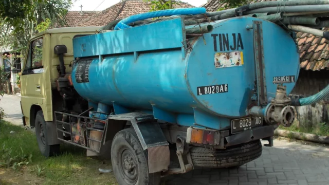 Truk tangki biru bertuliskan TINJA dengan selang sedot terparkir di jalan kampung