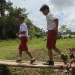 Dua murid sekolah dasar berseragam merah putih berjalan di titian kayu desa