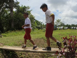 Dua murid sekolah dasar berseragam merah putih berjalan di titian kayu desa