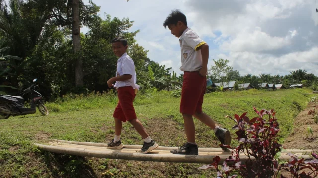 Dua murid sekolah dasar berseragam merah putih berjalan di titian kayu desa