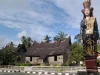 Patung perempuan berpakaian adat Minangkabau berdiri di tepi jalan dengan rumah gadang beratap gonjong di belakangnya