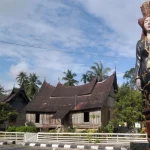 Patung perempuan berpakaian adat Minangkabau berdiri di tepi jalan dengan rumah gadang beratap gonjong di belakangnya