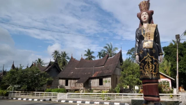 Patung perempuan berpakaian adat Minangkabau berdiri di tepi jalan dengan rumah gadang beratap gonjong di belakangnya