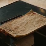 Ilustrasi lembar naskah tulisan tangan yang menguning di atas meja kayu gelap.