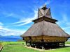 Rumah adat Karo beratap ijuk bertingkat berdiri di atas tiang kayu dengan latar danau dan langit biru.