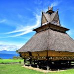 Rumah adat Karo beratap ijuk bertingkat berdiri di atas tiang kayu dengan latar danau dan langit biru.