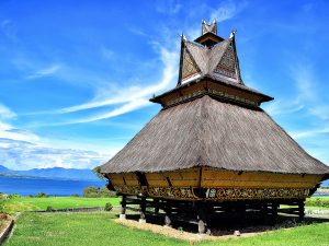 Rumah adat Karo beratap ijuk bertingkat berdiri di atas tiang kayu dengan latar danau dan langit biru.
