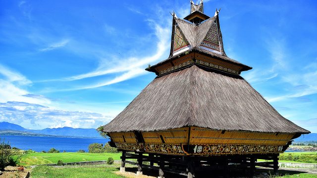 Rumah adat Karo beratap ijuk bertingkat berdiri di atas tiang kayu dengan latar danau dan langit biru.