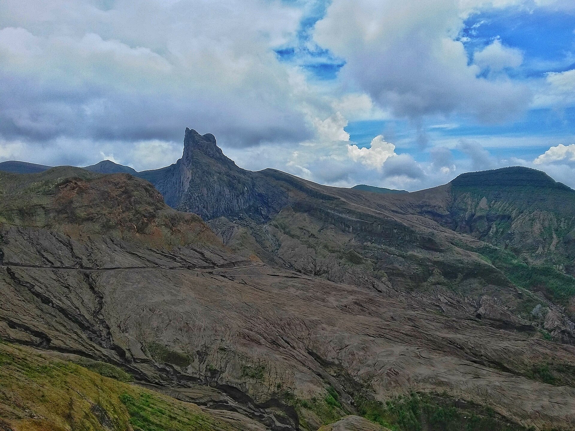 Puncak berbatu Gunung Kelud di Kediri menjulang di antara punggungan bukit hijau dan awan tebal.