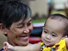 Nenek beruban tersenyum sambil menggendong cucu balita berbaju kuning di halaman rumah