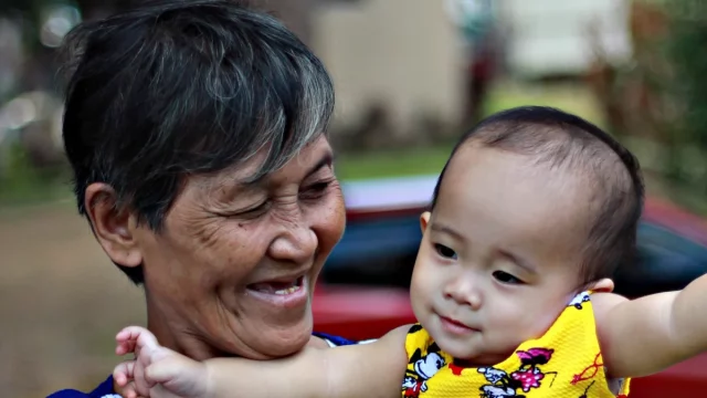 Nenek beruban tersenyum sambil menggendong cucu balita berbaju kuning di halaman rumah