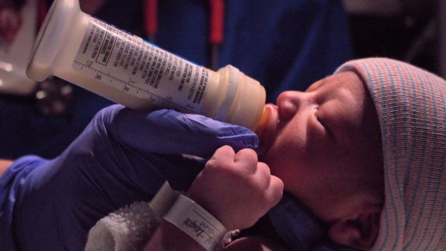 Ilustrasi bayi baru lahir disuapi susu botol oleh tangan bersarung tangan medis.