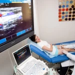 Seorang dokter melakukan pemeriksaan ultrasonografi Doppler pada pasien, dengan citra aliran darah berwarna tampil di layar besar
