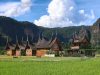 Ilustrasi rumah gadang beratap gonjong di tengah hamparan sawah dengan perbukitan di latar belakang.