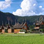 Ilustrasi rumah gadang beratap gonjong di tengah hamparan sawah dengan perbukitan di latar belakang.