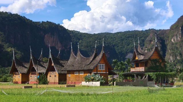 Ilustrasi rumah gadang beratap gonjong di tengah hamparan sawah dengan perbukitan di latar belakang.
