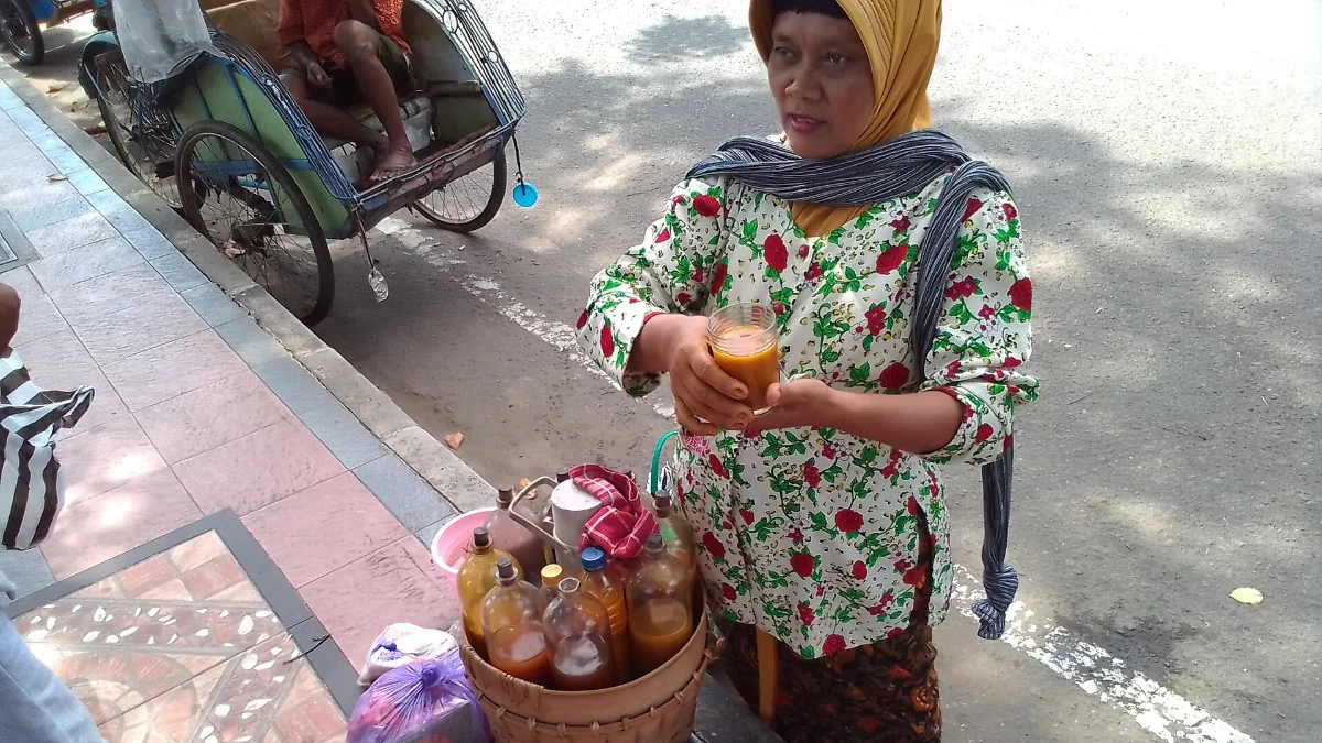 Botol Jamu yang Berpindah Arti dalam Abadi Nan Jaya 2 Seorang perempuan berdiri di tepi jalan memegang segelas jamu di samping keranjang botol