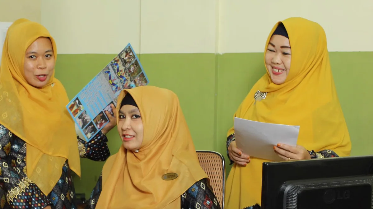 Tiga pekerja perempuan berkerudung kuning berbincang di depan komputer di sebuah ruang kantor