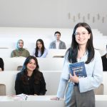 Ilustrasi seorang perempuan berdiri memegang buku di ruang kuliah bertingkat sementara sejumlah mahasiswa duduk di kursi di belakangnya.