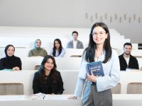 Ilustrasi seorang perempuan berdiri memegang buku di ruang kuliah bertingkat sementara sejumlah mahasiswa duduk di kursi di belakangnya.