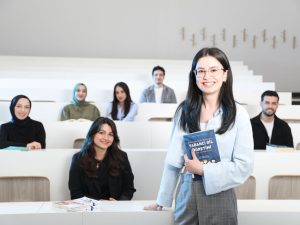 Ilustrasi seorang perempuan berdiri memegang buku di ruang kuliah bertingkat sementara sejumlah mahasiswa duduk di kursi di belakangnya.