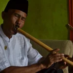 Seorang pemain meniup saluang darek sambil duduk di ruang tamu