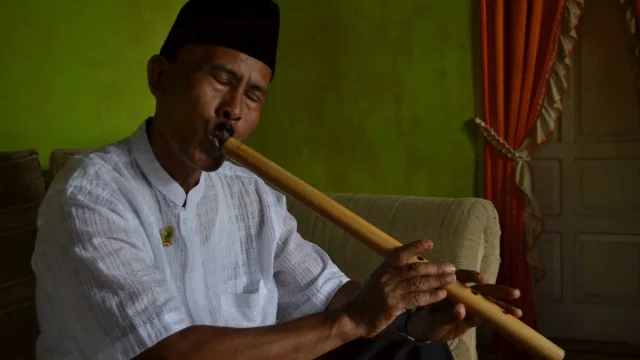 Seorang pemain meniup saluang darek sambil duduk di ruang tamu