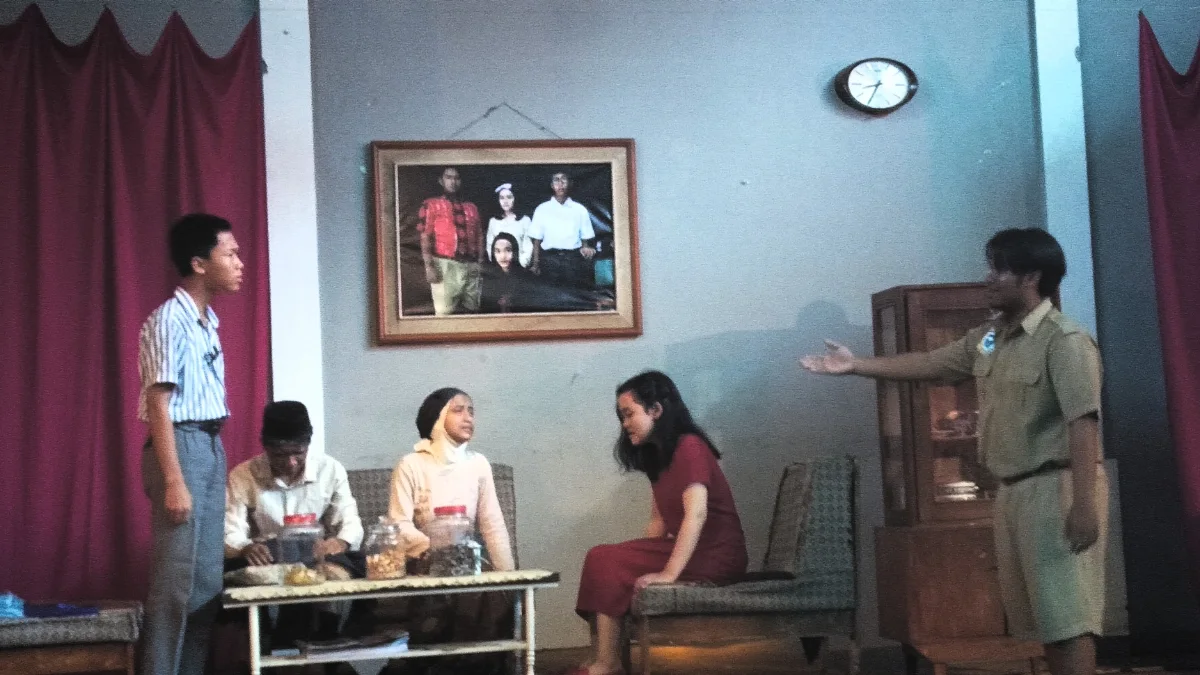 Seorang aktor berdiri sambil menunjuk ke arah tokoh lain dalam adegan panggung berlatar ruang tamu