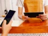 Pemilik kedai bercelemek memegang tablet kasir sementara pembeli menunjukkan ponsel untuk membayar