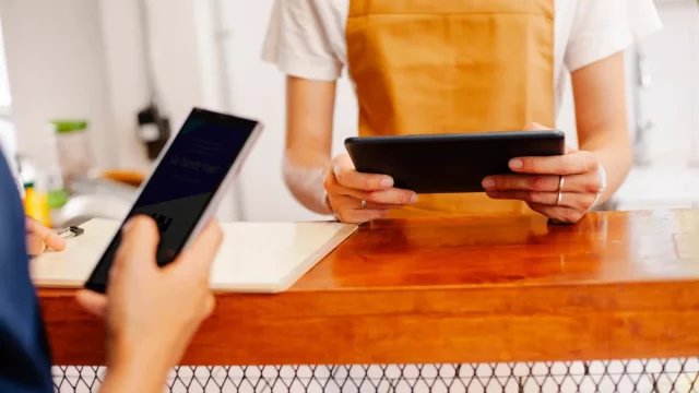 Pemilik kedai bercelemek memegang tablet kasir sementara pembeli menunjukkan ponsel untuk membayar