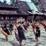 Penari Nias membawa tombak dan perisai dalam tarian perang di depan rumah adat