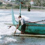 Ilustrasi nelayan berdiri di perahu tradisional saat mendorongnya ke arah gelombang.