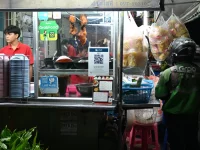 Pengemudi ojek daring berjaket hijau menunggu pesanan di depan kios makanan malam, Pattaya, Thailand