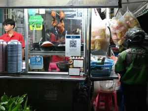 Pengemudi ojek daring berjaket hijau menunggu pesanan di depan kios makanan malam, Pattaya, Thailand