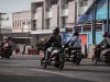 Beberapa pengendara sepeda motor berhelm melaju di jalan kota berlatar deretan ruko