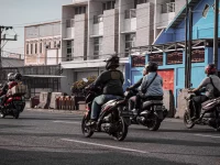 Beberapa pengendara sepeda motor berhelm melaju di jalan kota berlatar deretan ruko