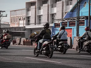 Beberapa pengendara sepeda motor berhelm melaju di jalan kota berlatar deretan ruko