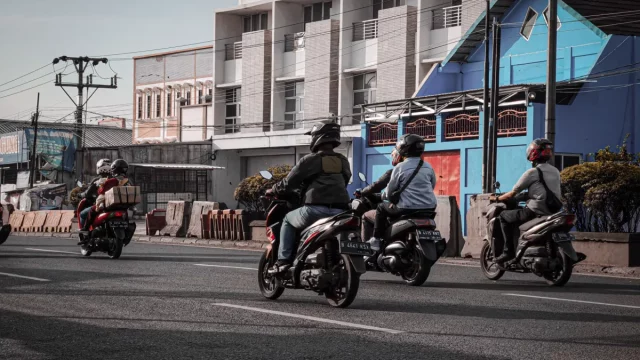 Beberapa pengendara sepeda motor berhelm melaju di jalan kota berlatar deretan ruko
