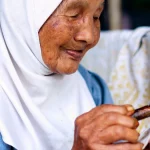 Perempuan lanjut usia berjilbab putih memegang canting di atas kain batik