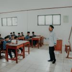 Ilustrasi guru berdiri di depan kelas berisi murid berseragam di bangku kayu.