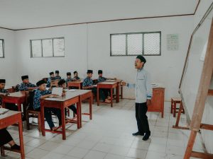 Ilustrasi guru berdiri di depan kelas berisi murid berseragam di bangku kayu.