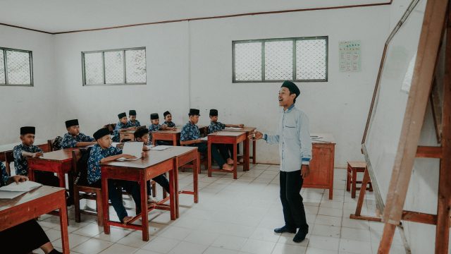 Ilustrasi guru berdiri di depan kelas berisi murid berseragam di bangku kayu.